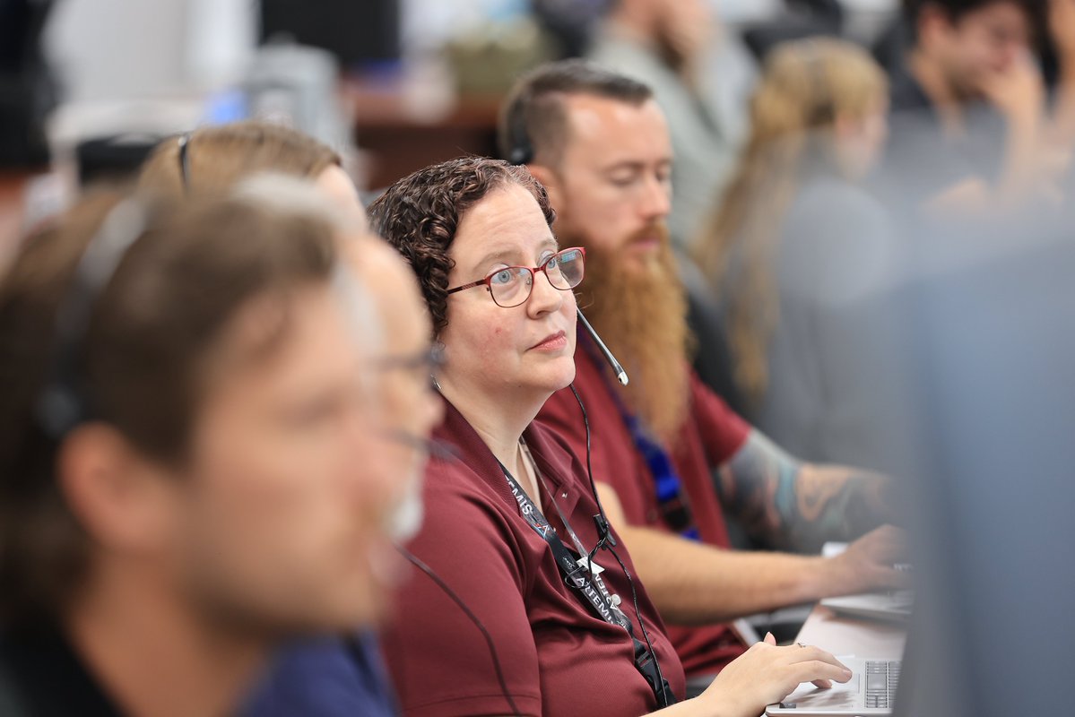 A woman wearing glasses and a red shirt and a lanyard with Artemis written on it, sits at a desk with a headset on surrounded by a group of others with headsets at NASA's Kennedy Space Center in Florida. Photo credit: NASA/Clayton Rougelot