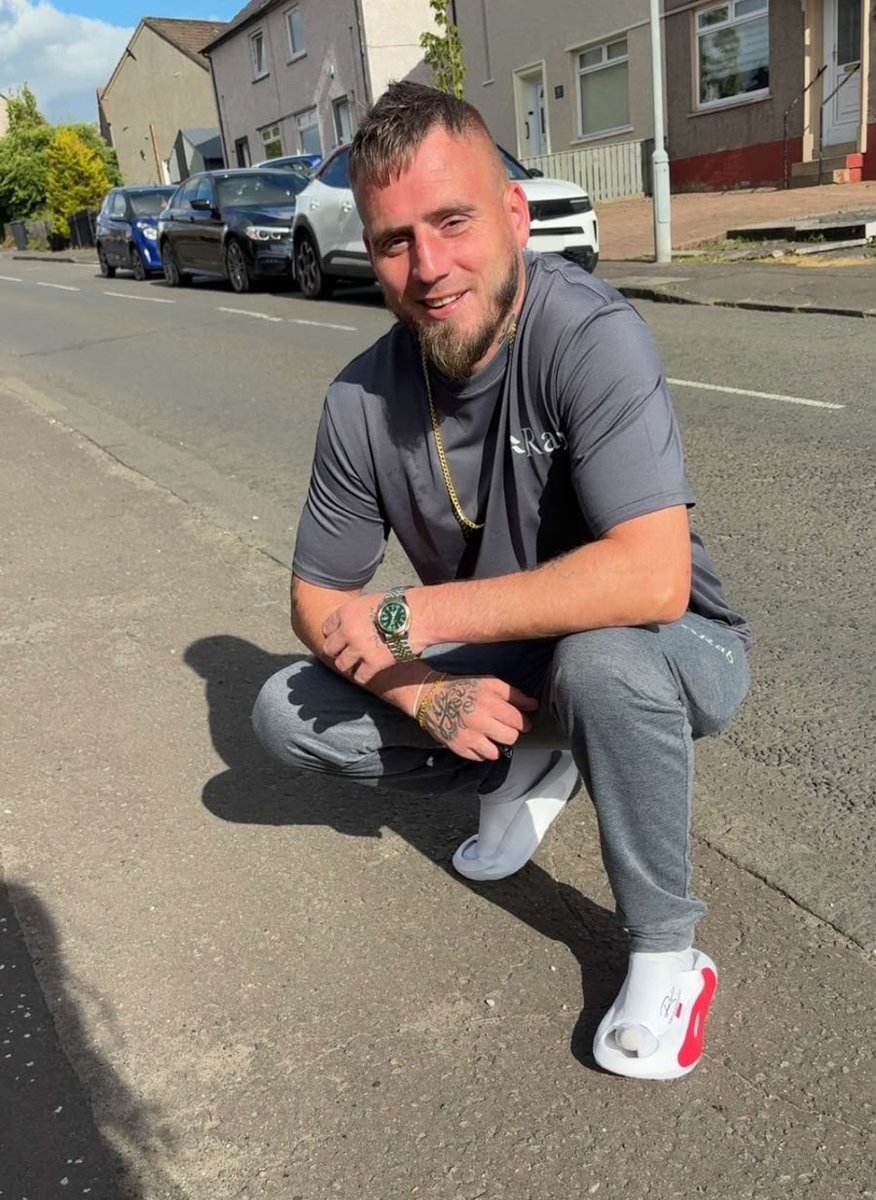 35-year-old man crouching in the street, smiling, wearing grey t-shirt and trousers with cars and houses in the background
