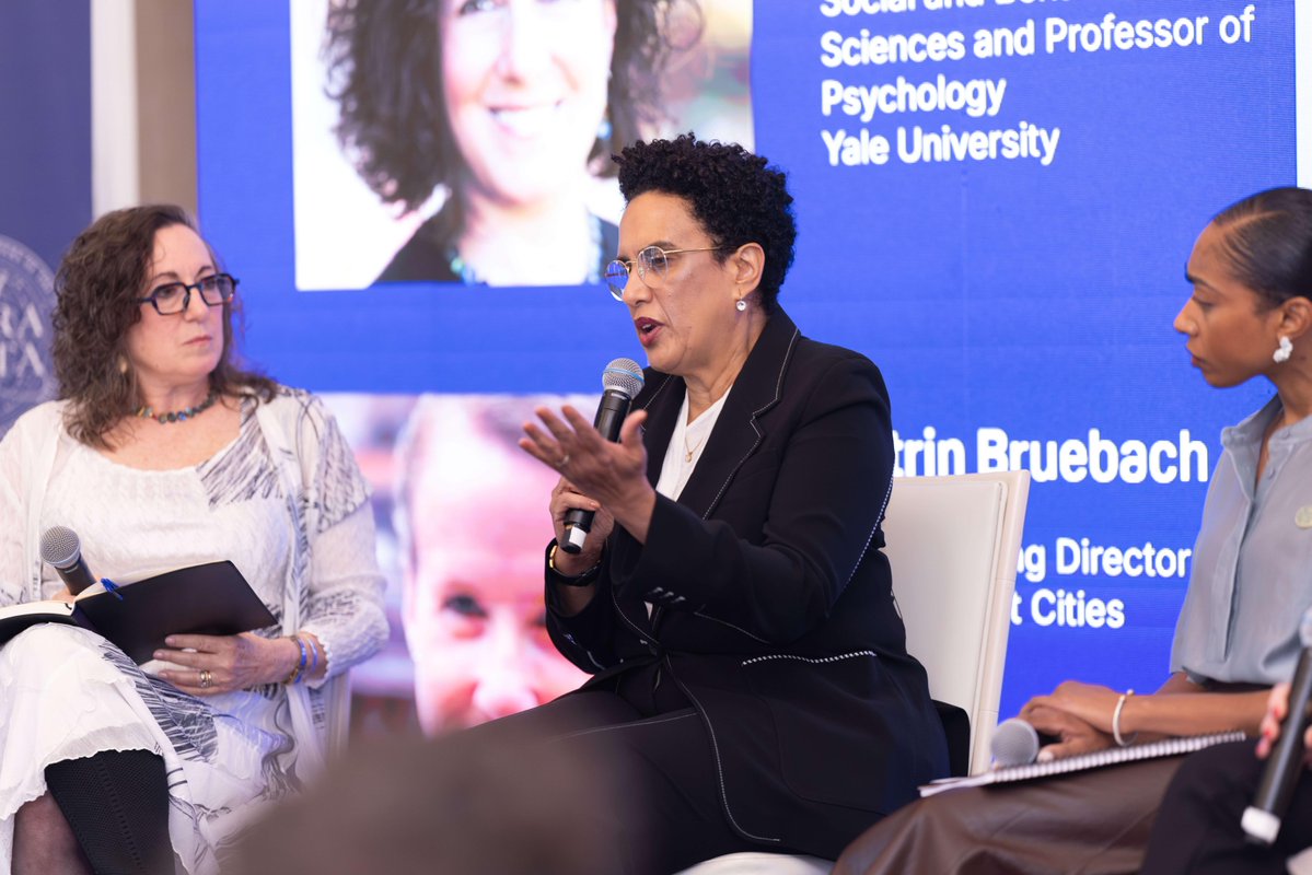 A panel discussion featuring three individuals seated on stage. The person in the center is speaking into a microphone. Behind them is a large screen displaying text, including the name "Yale University" and part of another name, "Bruebach." The individuals are engaged in conversation, with one person taking notes.