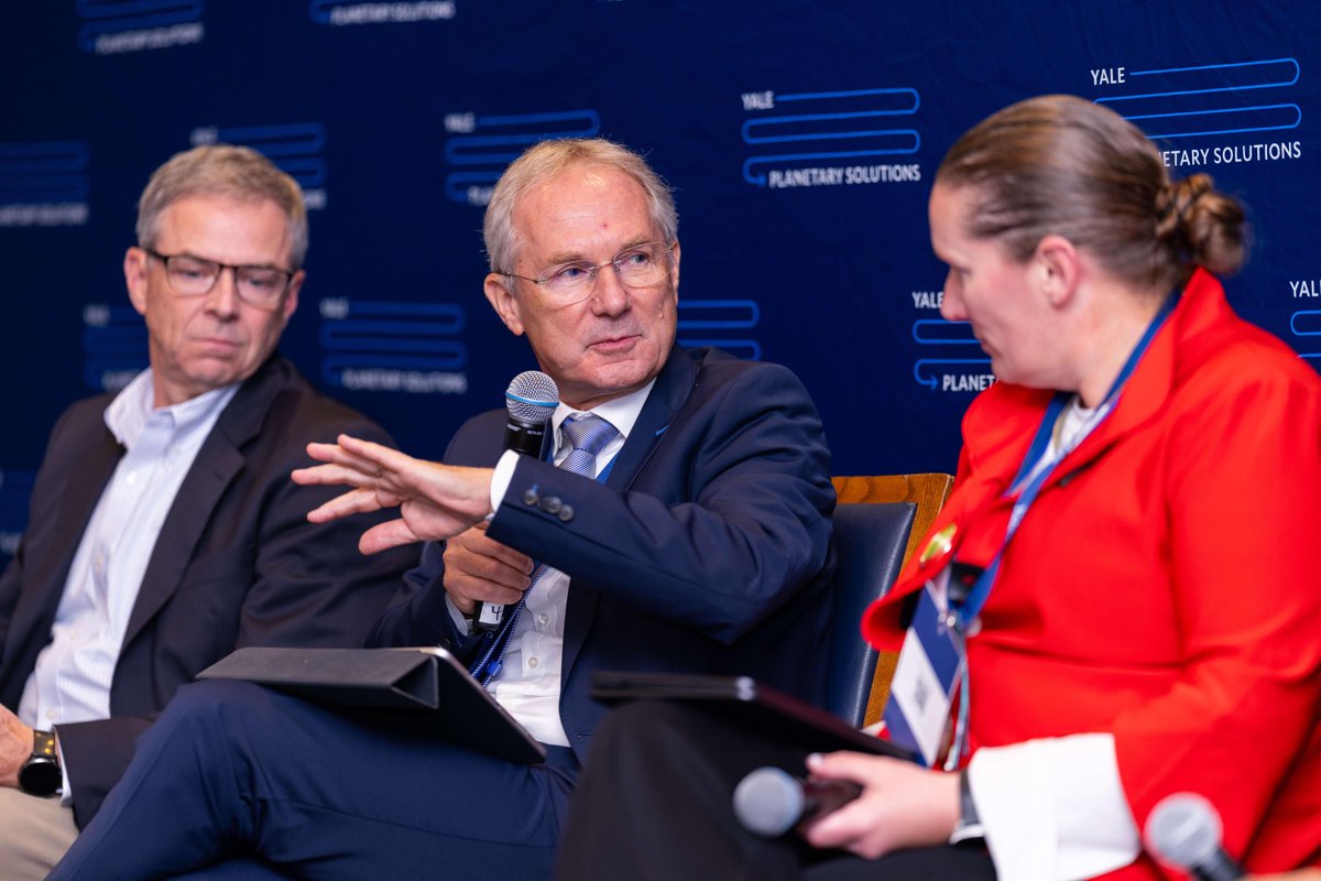 A group of three people engage in a discussion on a panel. They are seated in front of a backdrop with the Yale Planetary Solutions logo. One person is gesturing with a hand, while the others listen attentively, each holding a tablet.