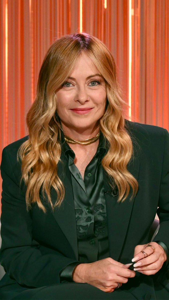 A blonde woman wearing a dark green blazer over a green patterned shirt and a gold choker necklace smiles at the camera against an orange backdrop.