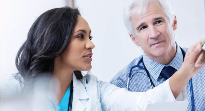 Two healthcare professionals in medical coats are engaged in discussion. One is gesturing towards something off-camera, while the other listens attentively. Stethoscopes are visible around their necks, suggesting they are doctors. The setting appears to be a medical office or hospital.