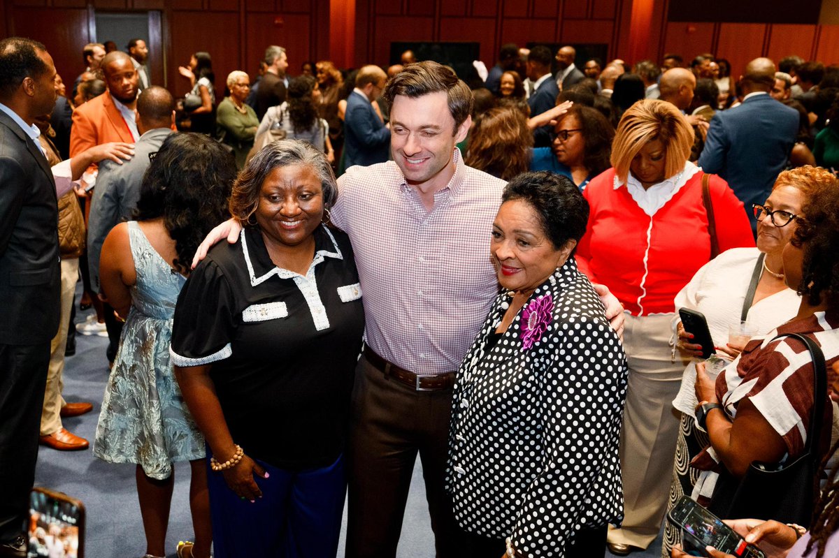 A man poses for a photo with two women at a crowded indoor gathering. People mingle in the background, while others nearby hold up phones to take pictures. There is no prominent readable text.
