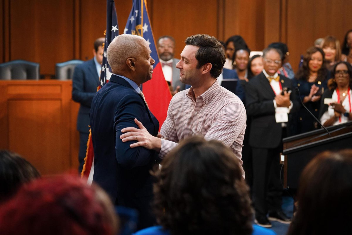 Two men greet each other warmly in a formal indoor venue while an audience applauds behind them. Flags and a lectern are visible; there is no prominent text.