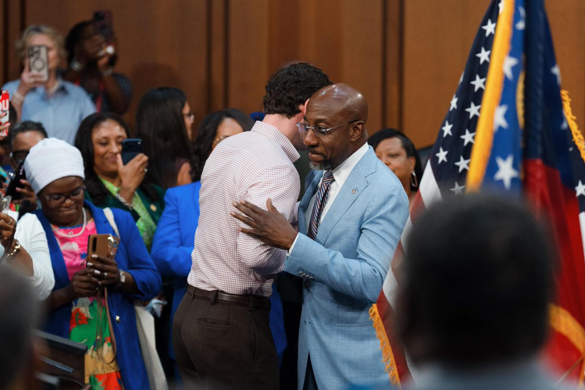 A man in a light blue suit greets another man in a crowded indoor gathering while people nearby watch and take photos. Several flags stand at the right side of the image. There is no prominent visible text.
