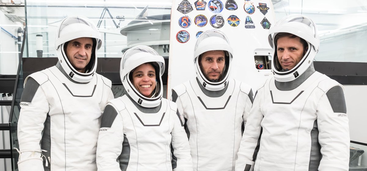 Four people wearing white SpaceX-style spacesuits stand together indoors, smiling or looking toward the camera. They are positioned in front of a wall displaying numerous colorful mission patches, with various spacecraft hardware visible in the background. Photo credit: SpaceX