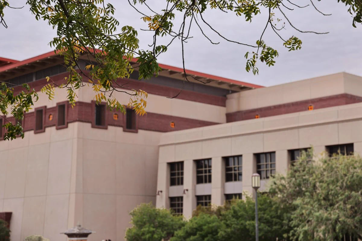 A large, multi-story building with a modern architectural style featuring beige and brick red walls on the UTEP campus. In the foreground, there are tree branches with green leaves, and some greenery at the base. The sky is overcast.
