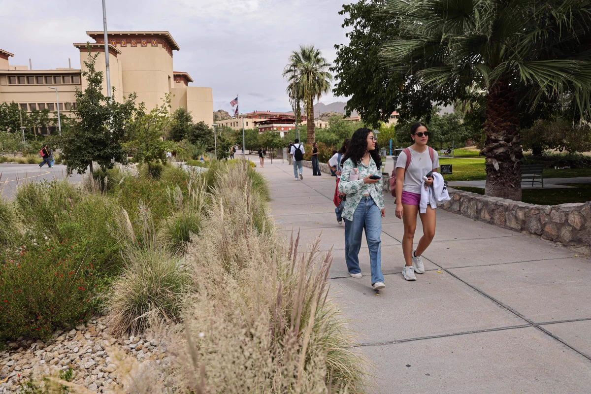 A sidewalk at the University of Texas at El Paso lined with greenery and palm trees. People are walking, and two individuals in the foreground are engaged in conversation. Buildings with Southwestern architecture are visible in the background.
