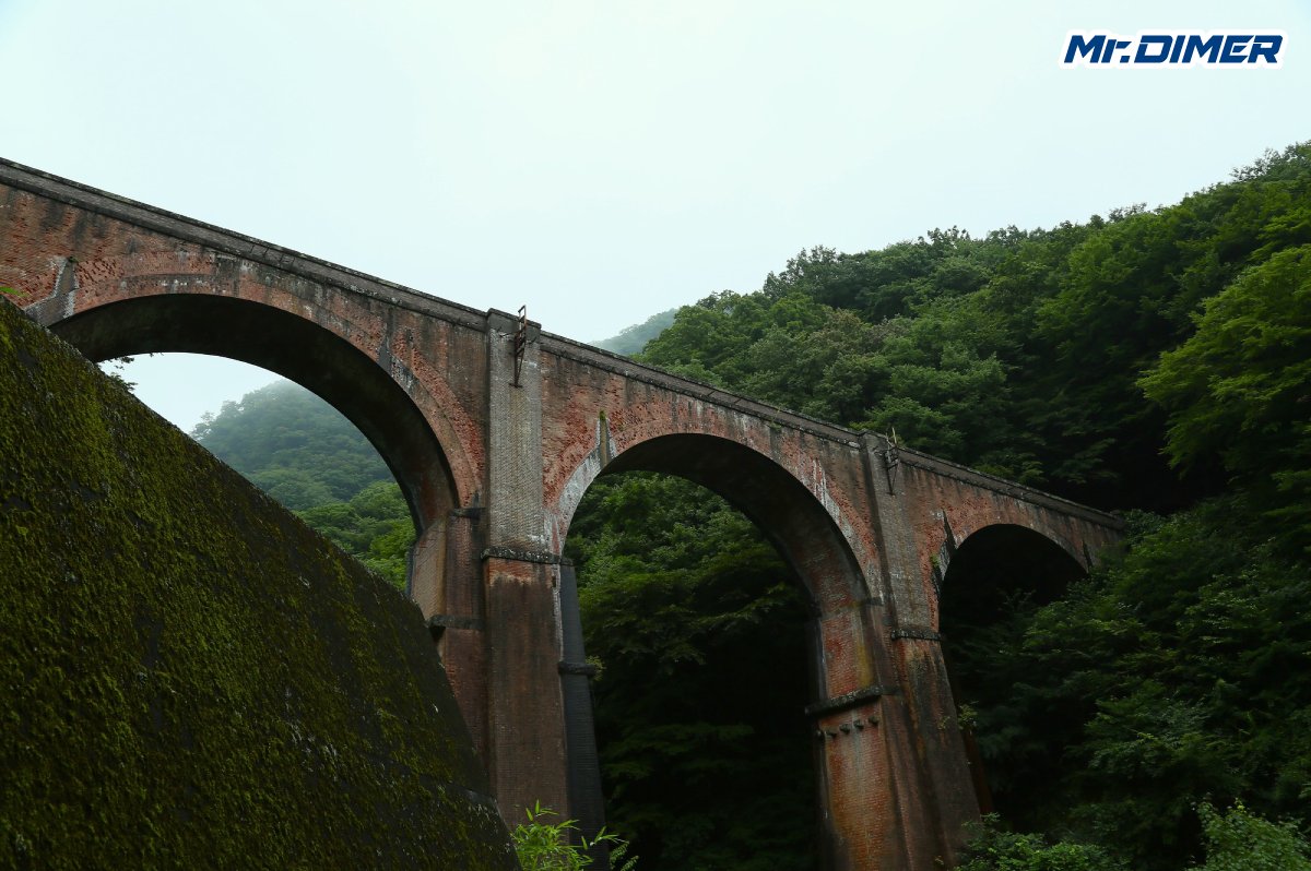 MrDIMER_X's tweet photo. 【なぜここに鉄道を通した】最大66.7‰、碓氷峠「横軽」104年の闘い

横川から軽井沢まで、わずか11kmあまり。
地図だけ見れば短い距離です。

でも、実際に碓氷峠へ行くと、
「よくここに鉄道を通したな」と思います。

山は深く、谷は急。
横川と軽井沢の標高差は500mを超え、
最大勾配は66.7‰。

鉄道にとっては、あまりにも急です。
それでも1893年、
横川と軽井沢の間に線路がつながりました。

ここを越えなければ、
東京側から延びてきた鉄道と、
長野、直江津方面へ続く鉄道が一本につながらない。

避けて通れない。
なら、越えるしかない。

そのために採用されたのがアプト式でした。
線路の中央に歯形のラックレールを置き、
機関車側の歯車を噛ませて登る。

今見れば特殊な仕組みですが、
当時はそこまでしなければ、
この峠を越えることができませんでした。

26本のトンネル。
18の橋梁。

今も残る「めがね橋」も、
最初から観光名所として造られたわけではありません。
列車を通すために必要だった。
ただ、それだけです。

ところが、
線路ができても苦労は終わりませんでした。
蒸気機関車で峠を登れば、
連続するトンネルの中に煙と煤がこもる。
時間もかかる。
輸送力にも限界がある。

そこで1912年には電化。
さらに戦後、
列車が増えれば、
今度はアプト式そのものが輸送の壁になっていきました。

そして1963年。

国鉄はアプト式をやめ、
新線による粘着運転へ切り替えます。

ここで登場したのがEF63でした。
碓氷峠専用。
ただ列車を押すための機関車ではありません。

66.7‰を登らせ、
そして下り坂では、
何があっても列車を止める。

横川側に連結されたEF63は、
この峠を越える列車の命を預かりました。

「あさま」が横川に着く。
EF63が近づいてくる。
連結する。
確認する。
そして、峠へ向かう。

特急も、急行も、普通列車も、
ここを越える列車は、
横川でひと手間かけなければ先へ進めませんでした。
今の感覚なら、非効率です。
でも、それが横軽でした。

ホームではEF63の連結を待ち、
釜めしを買う乗客がいて、
やがて列車は軽井沢へ向けて動き出す。

そんな光景が、
何十年も当たり前のように続いていました。

そして1997年9月30日。
その日常が終わります。

最後の日も、
横川ではいつものようにEF63が連結され、
列車は峠へ向かいました。
昨日までと同じ動き。
昨日までと同じ音。

でも、その一つひとつが最後でした。

多くの人が横川に集まり、
見慣れたはずの連結作業を見つめ、
峠へ消えていく列車を見送りました。

翌日には長野新幹線が開業。
信越本線・横川―軽井沢間は廃止されました。

1893年から104年。

最初は歯車で山に食らいつき、
次に電気の力を使い、
最後はEF63が列車を押し上げた。
方式は何度も変わりました。

でも、
横軽がやってきたことは最初から最後まで同じです。
横川と軽井沢の間にある、
この険しい山を越える。
そのためだけに、
鉄道は何度も姿を変えました。

普通なら、
廃線になれば痕跡は少しずつ消えていきます。
線路が剥がされ、建物がなくなり、
車両も姿を消していく。
でも横川は違いました。

めがね橋。
旧丸山変電所。
残された線路。
そしてEF63。

さらに旧横川運転区の跡地には、
碓氷峠鉄道文化むらが残りました。

横軽の歴史は、
車両だけ残しても伝わりません。

なぜEF63が必要だったのか。
なぜ横川で連結したのか。
なぜこの町にこれほど大きな鉄道施設があったのか。

その理由まで含めて残っている。
ただ昔の車両を並べただけではない。

横軽で実際に使われたEF63を、
今も動かしている。
講習を受ければ、
本物のEF63を自分で運転することもできる。

廃止から長い時間が経った今も、
横川へ行けば、
あの峠を越えた機関車の音がする。
横軽はもうありません。

列車が横川から軽井沢へ登っていくこともありません。
それでも、
橋も、変電所も、線路も、機関車も残った。

だから今でも、
104年間ここで何が行われていたのかを、
現物を見ながら知ることができます。

1997年9月30日。

あの日、
横川から軽井沢へ向かう列車は止まりました。

けれど、
104年間この峠と向き合ってきた鉄道の記憶まで、
一緒に消えたわけではありません。

最大66.7‰。
なぜ、そこまでしてこの峠を越えなければならなかったのか。
なぜ、専用の機関車まで造ったのか。

そして、
なぜ横軽は104年間も走り続けることができたのか。

9月30日。

横軽が止まったこの日に、
その答えをもう一度辿ります。

次回の記事は、
66.7‰。

碓氷峠は、
なぜこれほどまでの急勾配になったのか。
104年間の歴史を少しずつ辿ってみましょう。

（マイミー）