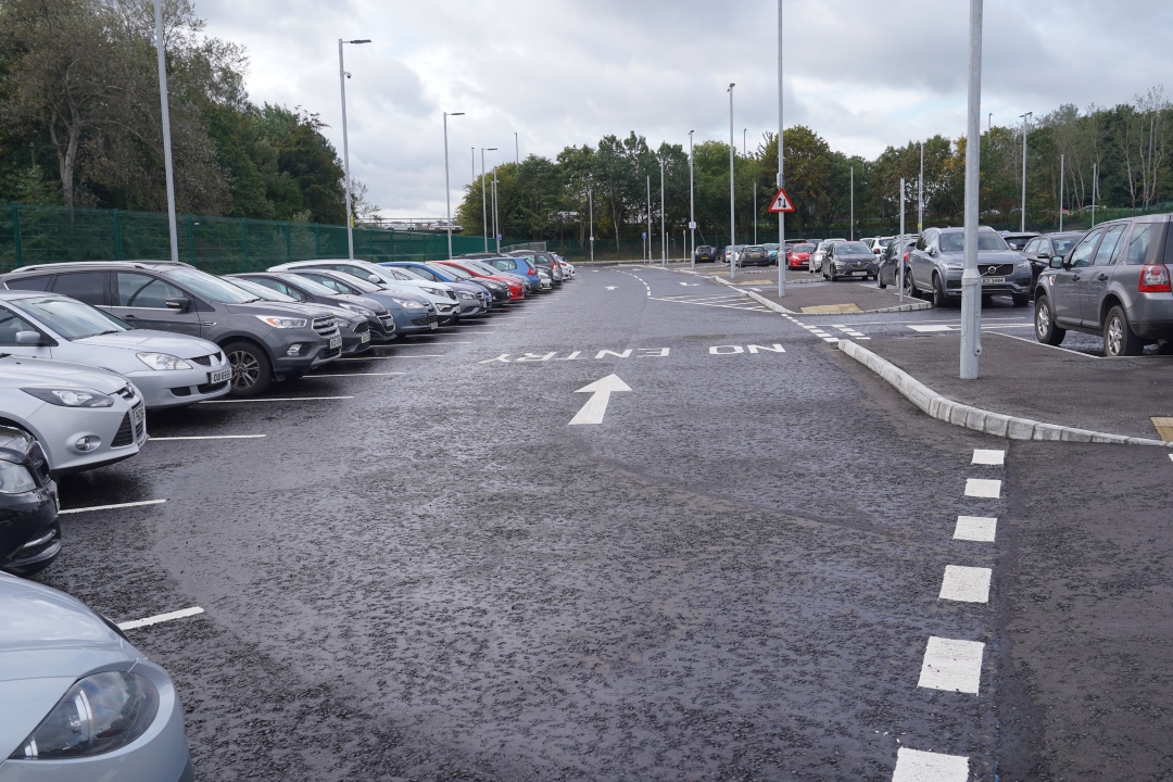 A car park with numerous cars parked in rows on the left and right. A white arrow points forward on the dark asphalt, with \"NO ENTRY\" written backward behind it. Trees and tall lampposts line the background. A red triangular warning sign with two arrows points up and down in the distance.