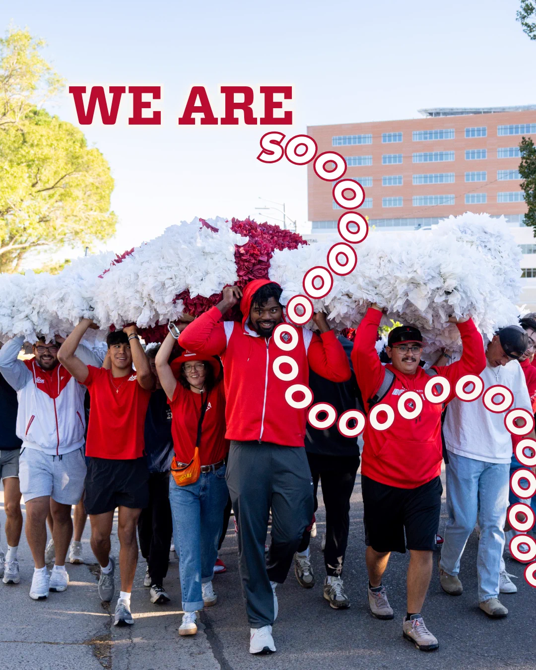 A group of students in red and white attire is parading, carrying the Aggie effigy. The words "WE ARE SOOOO" are prominently displayed above them in bold red letters. A building and trees are visible in the background.