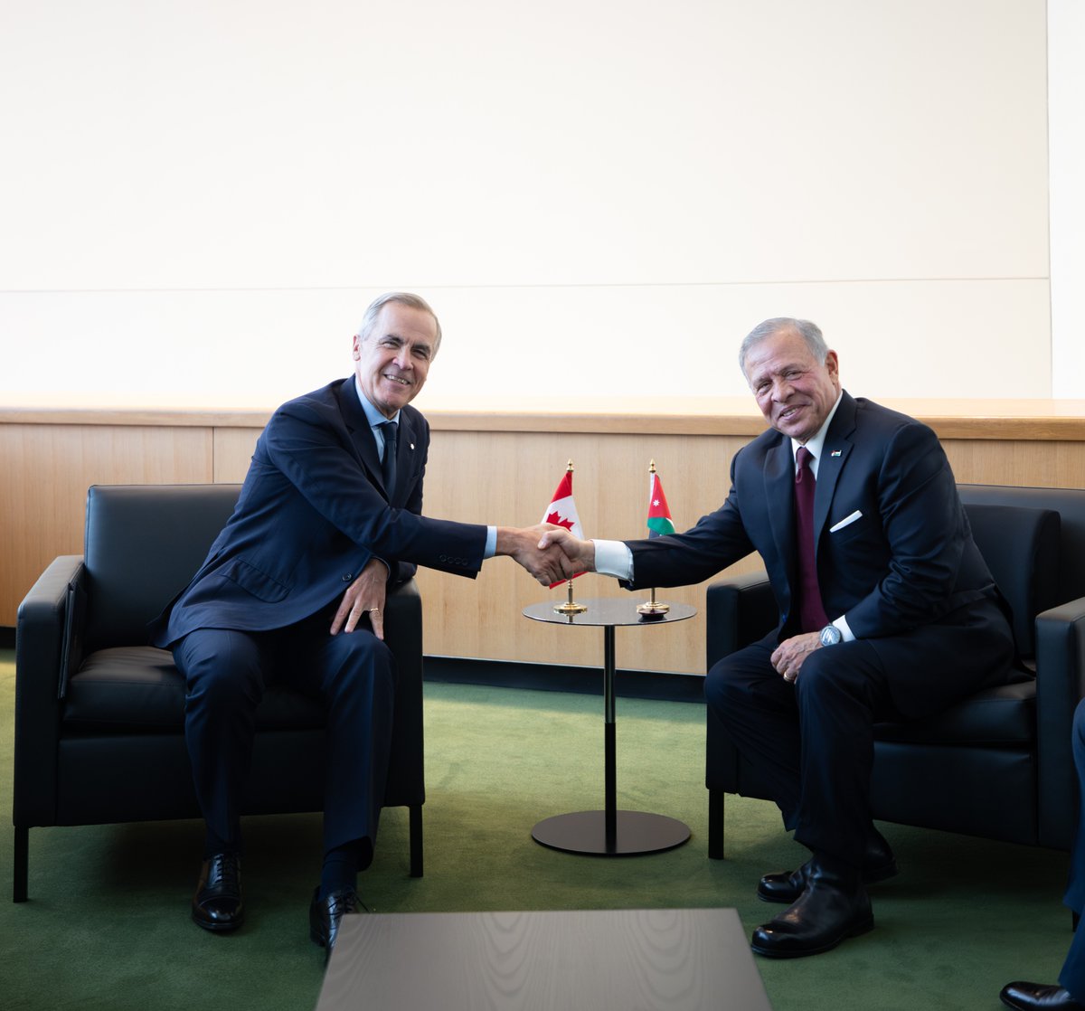 Prime Minister Mark Carney and His Majesty King Abdullah II of Jordan shake hands and smile. Small flags of Canada and Jordan are on a table between them.