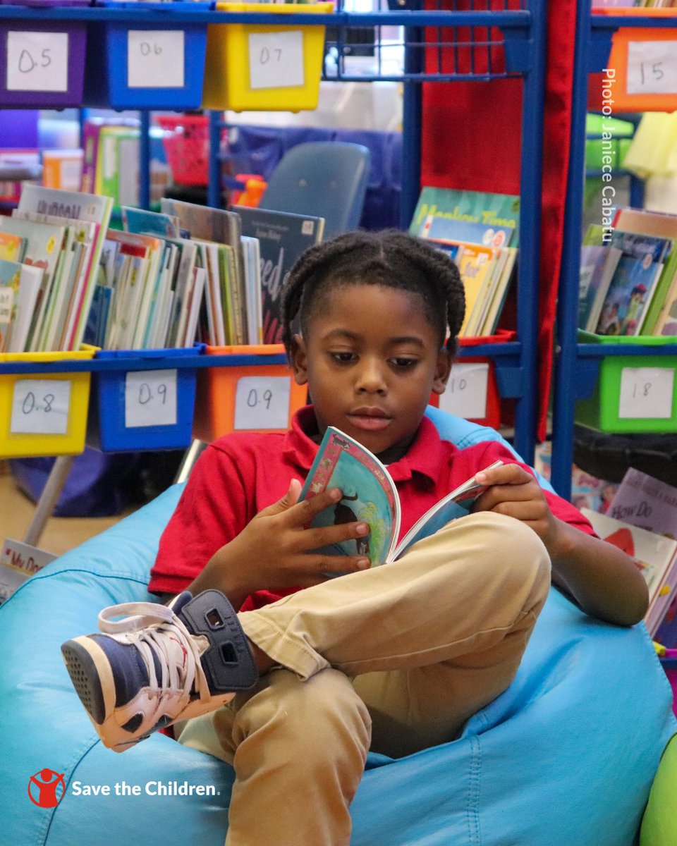 A child is sitting on a blue bean bag chair, reading a book in a library. Bookshelves filled with colorful books are in the background. The "Save the Children" logo is visible in the bottom left corner.