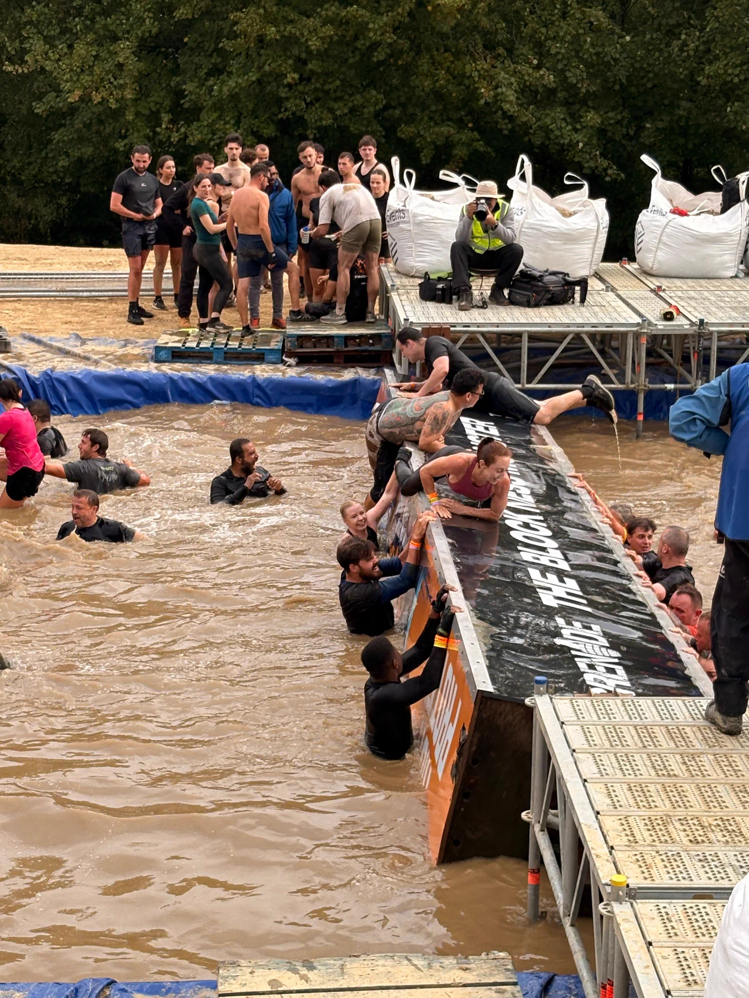 Participants helping each other climb a muddy obstacle during an outdoor endurance race in a wooded area.