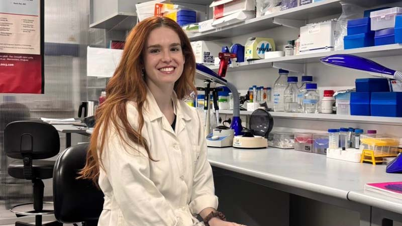 Mujer con bata blanca sonriendo, sentada en un laboratorio junto a una mesa con equipos, frascos y reactivos.