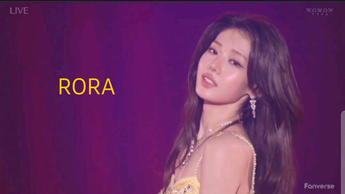 A young woman labeled "RORA" posing against a purple backdrop during a WOWOW Prime live broadcast, wearing a yellow outfit with pearl and chain accessories.