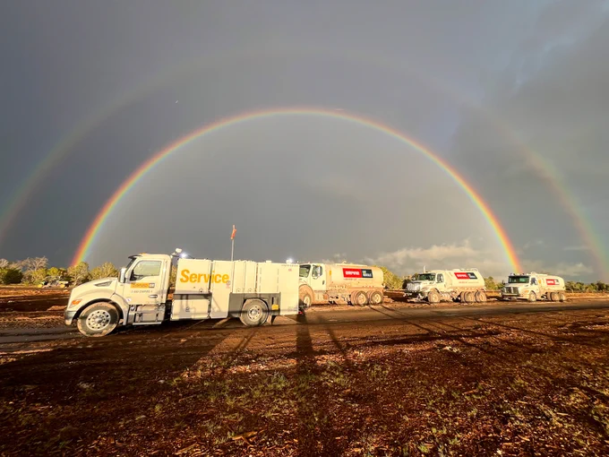 A fleet of trucks with visible Empire Cat Rentals stands on a muddy terrain under a bright double rainbow. The trucks are lined up, facing right, with the words "Service,". The sky is partly cloudy, and the ground is wet, reflecting the light from the rainbow.
