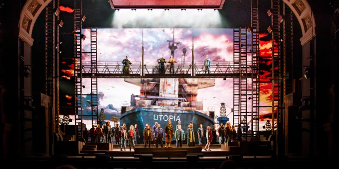 A large stage production featuring a diverse cast. The backdrop displays a large digital image of a ship with the word "UTOPIA" on it. The scene includes a blend of vibrant costumes and dynamic lighting, suggesting a dramatic or musical performance.