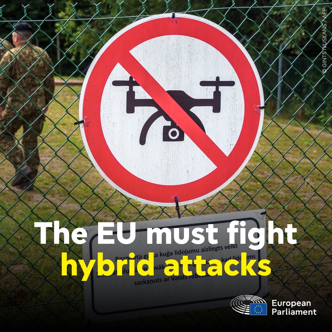 "The EU must fight hybrid attacks" reads the text on this photo of a sign on a chain-link fence. The sign prohibits drone flights, featuring a silhouette of a drone with a red circle around it and a red line across it. Soldiers are visible in the background on the other side of the fence.