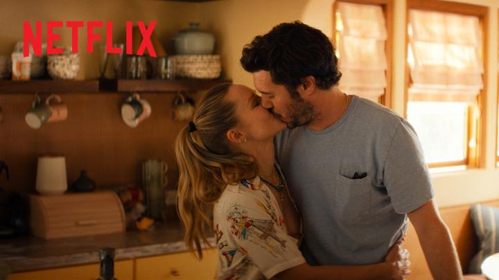 The image shows a couple sharing a kiss in a sunlit kitchen, with the "NETFLIX" logo visible in the upper left corner, promoting the romantic series "Nobody Wants This." The intimate moment between the blonde woman and bearded man reflects the show's central romance, aligning with the caption's sentiment about their relationship being "perfect because it's us" ahead of Season 3's October 22 release.