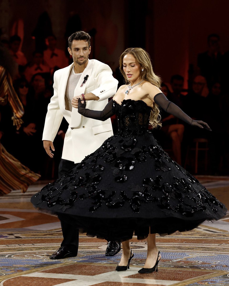popbuzzz_'s tweet photo. Jennifer Lopez stuns at the Vogue World 2026: Milano in the Galleria Vittorio Emanuele II wearing a strapless black Dolce & Gabbana tulle ballgown with floral sequin appliqués, sheer opera gloves, pointed slingbacks, and a standout 74-carat turquoise pendant necklace.

The star was pulled from the front row by Italian tennis player Lorenzo Musetti for a surprise runway dance moment evoking classic Italian cinema like La Dolce Vita, highlighting the event’s celebration of Made in Italy craftsmanship during Milan Fashion Week.