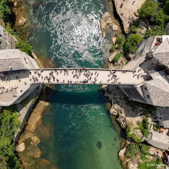 Aerial view of a bridge spanning a turquoise river. People are walking across the bridge, and lush greenery surrounds the area.