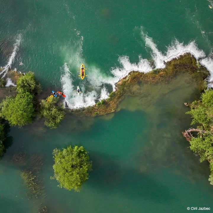 Aerial view of two kayakers navigating through a narrow channel of clear, green water, flanked by trees and shallow rapids.