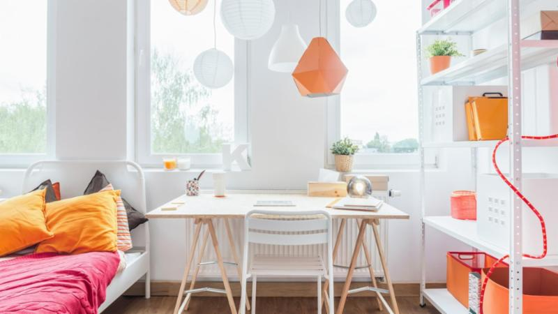 Dormitorio luminoso con escritorio junto a ventanas, cama roja/naranja, lámparas colgantes y estantería con cajas naranjas.