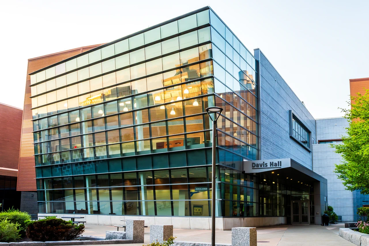 Modern university building, Davis Hall, features a striking glass facade reflecting warm golden light, alongside reddish-brown and grey paneling. A paved walkway leads to the entrance, with landscaping and outdoor seating areas.