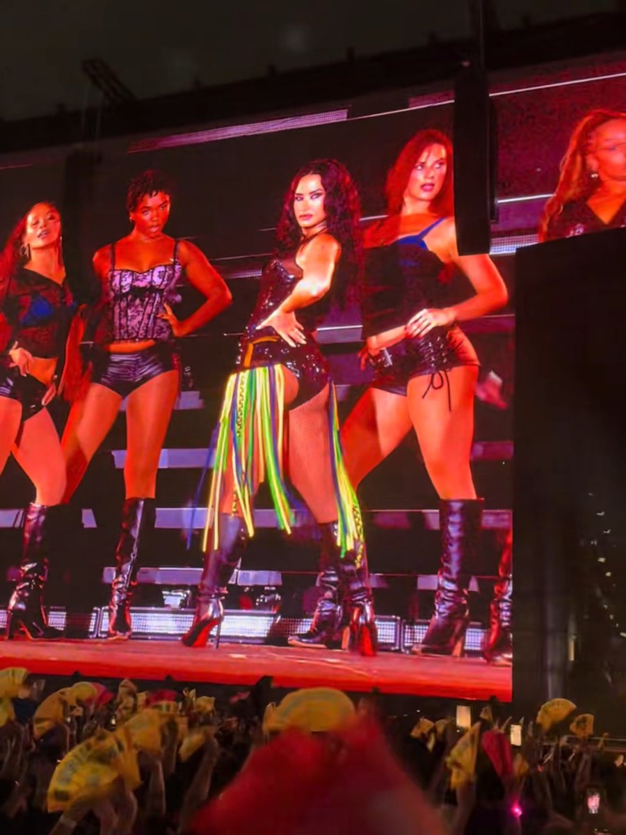 A concert performance featuring a woman in a black outfit with colorful fringe dancing with backup dancers on stage under red lighting.