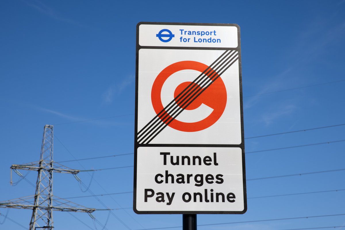 A white rectangular sign against a blue sky. It displays the Transport for London logo and text, an orange circle with a 'C' crossed by black lines, and 'Tunnel charges Pay online'. Part of an electricity pylon is visible left.