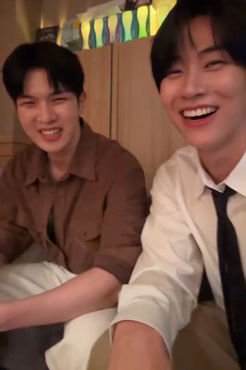 Two young men with dark hair sit side by side in what appears to be a casual indoor setting, both grinning brightly at the camera. The man on the right wears a white shirt with a dark tie and laughs openly with his eyes crinkled, while the man on the left in a brown button-up shirt smiles with a slightly scrunched, playful expression, capturing the carefree, giddy energy described in the caption.