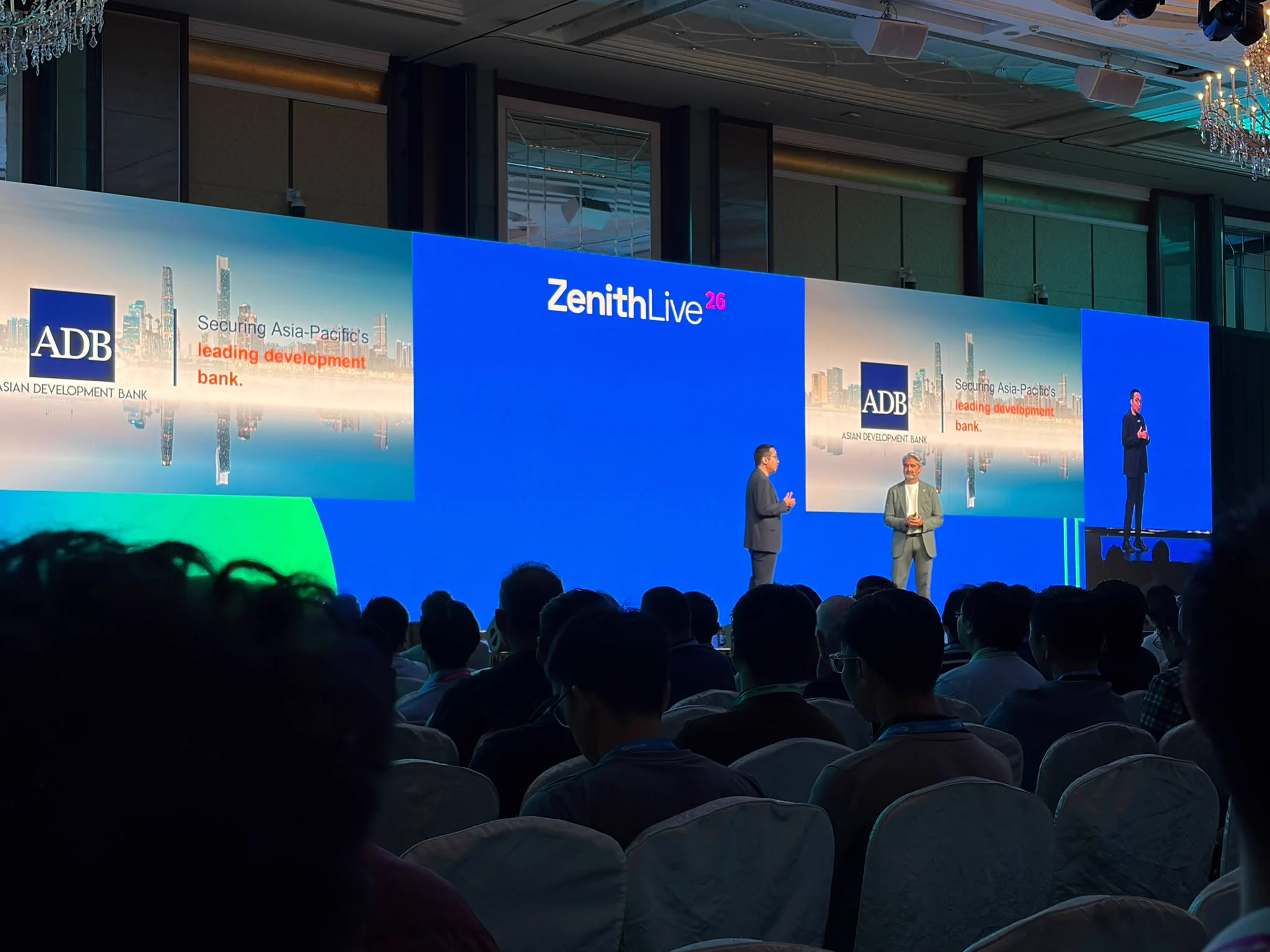 A conference setting with a large stage, featuring two speakers addressing the audience. Behind them, a screen displays the logos of Asian Development Bank (ADB) and Zenith Live. The audience is seated facing the stage.