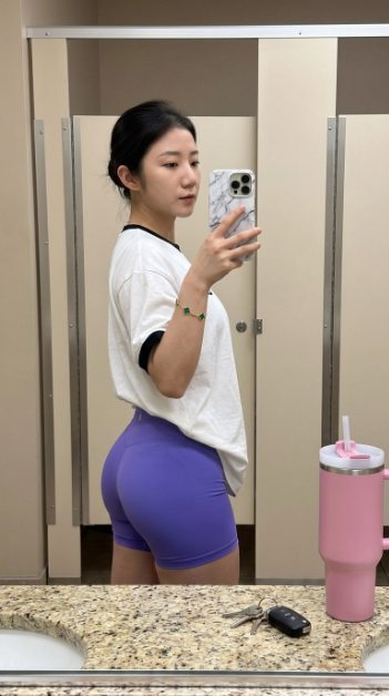 aisima2046's tweet photo. 分享一下这个的提示词：
Json格式，效果拉满
Prompt :-
{
  "image_analysis": {
    "composition": {
      "aspect_ratio": "9:16 (Vertical Portrait)",
      "shot_type": "Mirror selfie, three-quarter side profile",
      "lighting": "Soft, diffused indoor overhead lighting, neutral color temperature",
      "focus": "Sharp focus on the subject with a natural depth of field"
    },
    "subject_details": {
      "use_reference_image": true,
      "override": false,
      "body_details": {
        "pose": "Standing side-on, twisting slightly toward the mirror, holding phone at chest level",
        "physique": "Athletic, curvy build"
      }
    },
    "attire_and_accessories": {
      "top": "Oversized white cotton ringer t-shirt with black ribbed cuffs",
      "bottom": "High-waisted, form-fitting athletic compression shorts in vibrant violet-purple",
      "jewelry": "Gold delicate chain bracelet featuring five green malachite four-leaf clover motifs (Van Cleef style)",
      "phone_case": "iPhone with a white and grey marble-patterned hardshell case, triple-lens camera visible"
    },
    "environment_and_objects": {
      "setting": "Minimalist fitting room or locker room stall",
      "background_walls": "Neutral beige/cream panels with vertical silver metal separation strips",
      "countertop": "Speckled granite or polished stone surface with tan and grey flecks",
      "objects_on_counter": [
        {
          "item": "Insulated tumbler",
          "details": "Large matte pink 'Stanley' style cup with a lid and reusable straw"
        },
        {
          "item": "Keys",
          "details": "Silver metal keys attached to a black electronic car key fob/remote"
        }
      ]
    }
  }
}