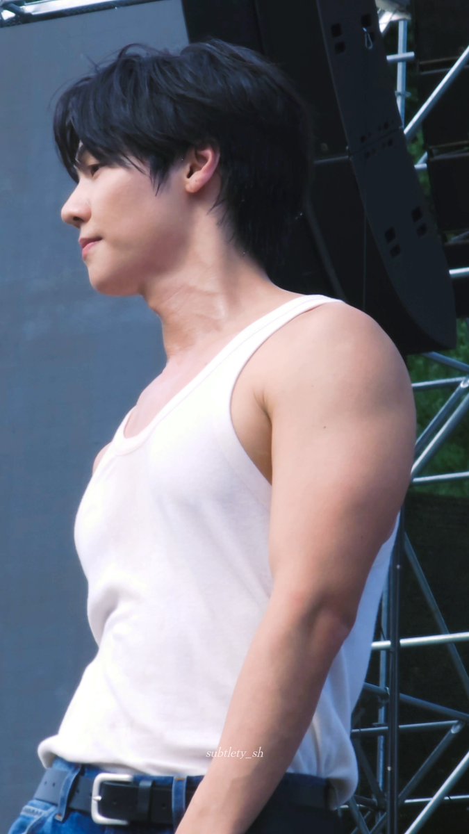 A side profile shot of a young man with black hair wearing a white tank top and blue jeans, standing near a metal scaffolding structure, with a 'subtlety_sh' watermark.