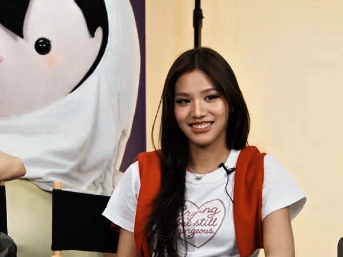 A woman with long dark hair, wearing a white t-shirt with a heart design and an orange vest, smiles while sitting in a chair with a cartoon character backdrop visible behind her.