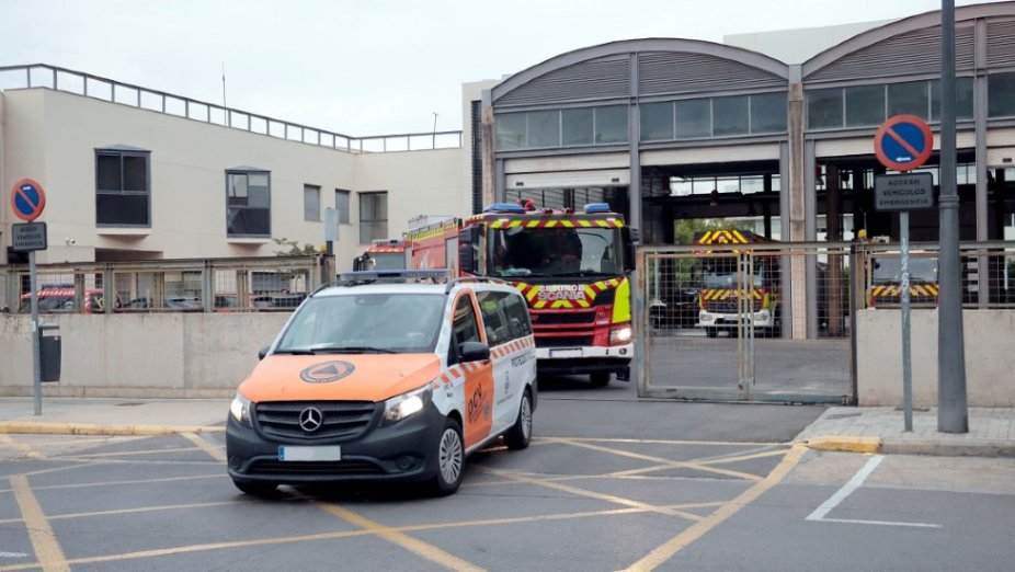 Vehículo de protección civil naranja al frente y camión de bomberos saliendo de un parque de bomberos urbano.