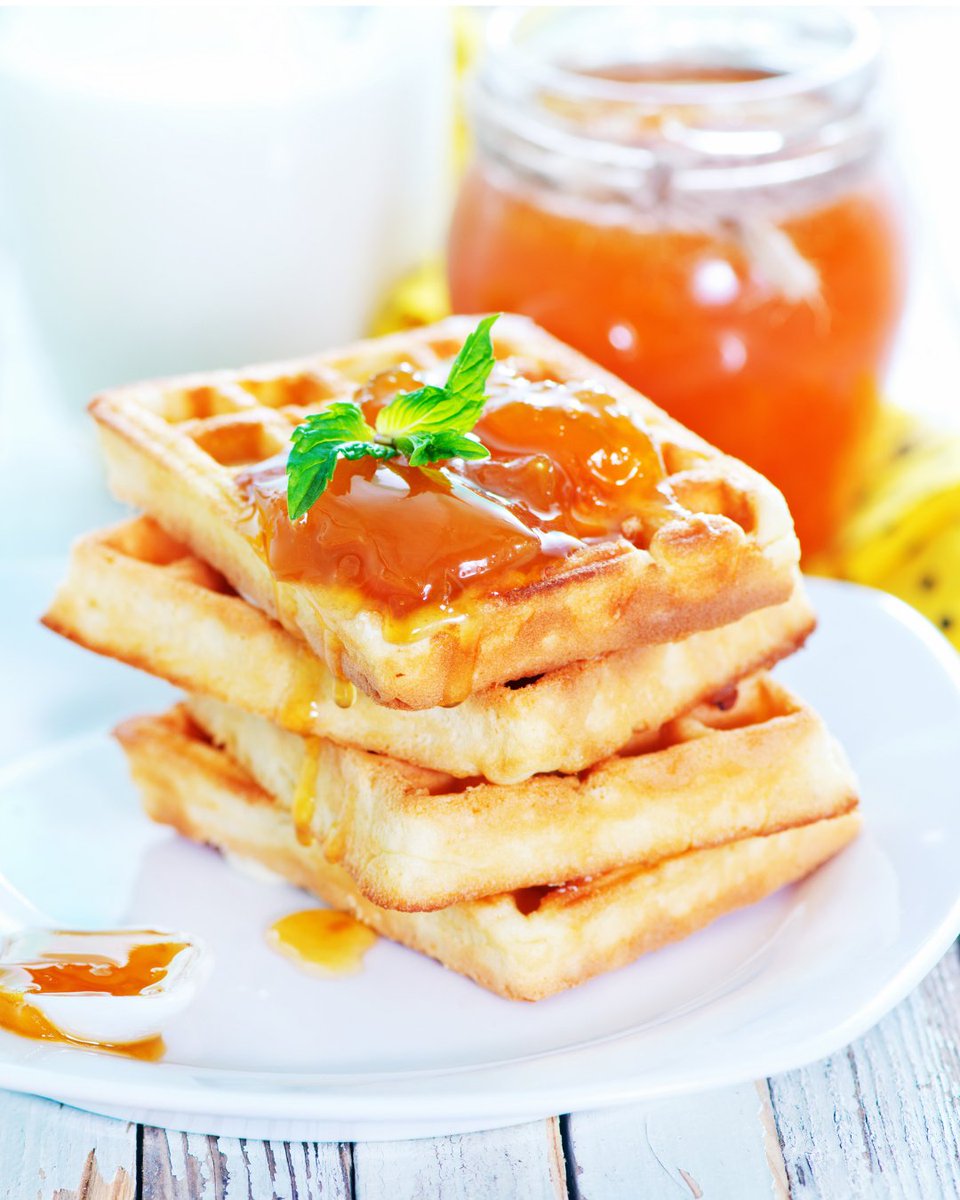 A stack of three golden waffles topped with a dollop of mandarin jam and a sprig of mint on a white plate. A jar of mandarin jam in the background.