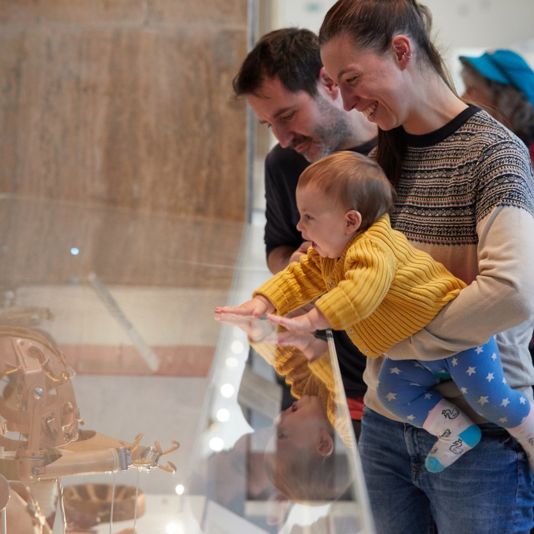 A person holds a baby in front of a museum display, both showing interest in a wooden artifact. Another individual stands nearby, observing.