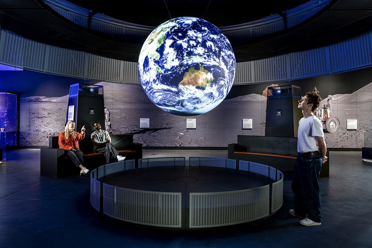 A group of people observe a large, suspended 3D model of Earth inside a modern exhibition space.