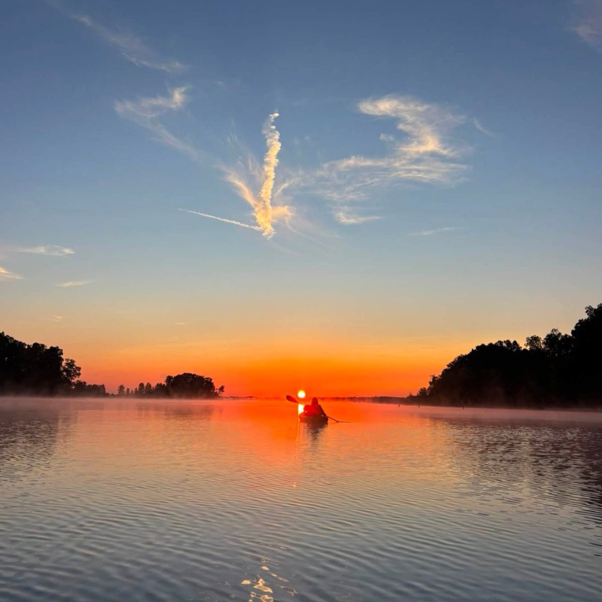 A person kayaks on a calm river during a vibrant sunset, with the sun setting near the horizon. The sky transitions from orange near the horizon to deep blue at the top, with a few scattered clouds. Trees line the riverbanks, and a mist hovers over the water.