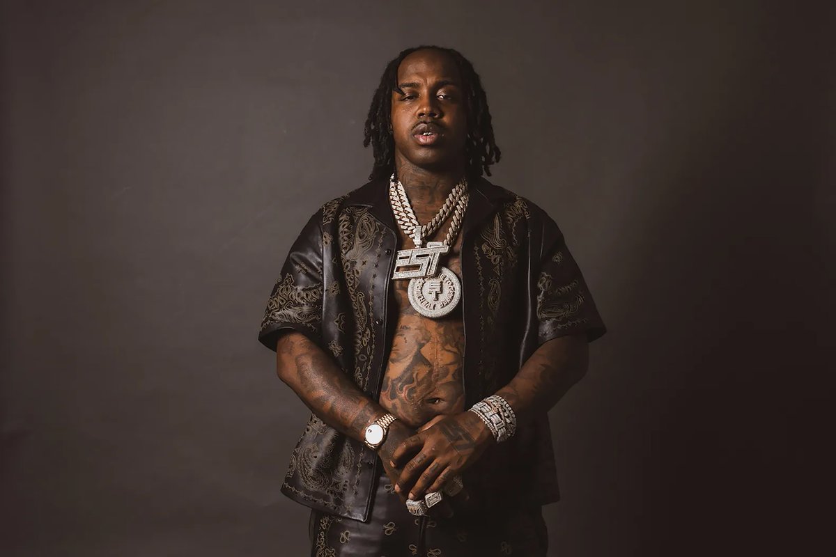 Man with long dreadlocks wearing an ornately patterned black leather short-sleeve jacket over a black tank top, displaying multiple diamond-encrusted chains, a large pendant necklace, diamond bracelets, and a watch against a neutral gray background.