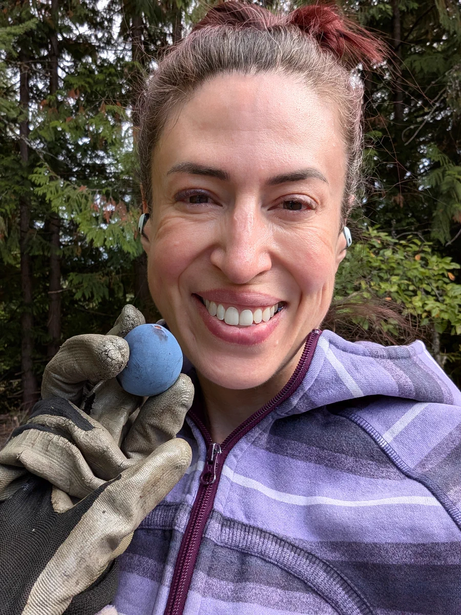 Smiling woman outdoors in front of evergreen trees, wearing a purple striped jacket and gloves, holding a blue berry or small fruit up beside her face.