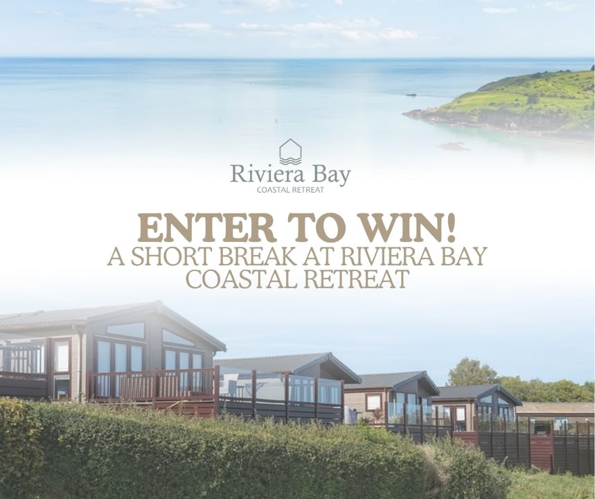 Modern coastal lodges sit on a grassy hillside overlooking calm blue sea and a green headland. The sky is clear and bright. Text on the image reads “Riviera Bay Coastal Retreat – Enter to win! A short break at Riviera Bay Coastal Retreat.”