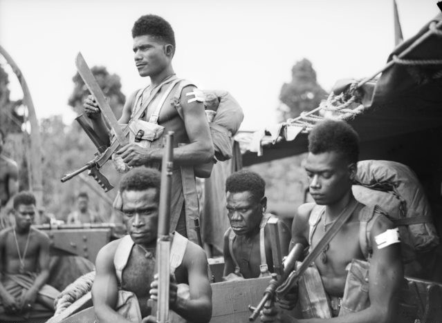 Soldiers holding rifles and wearing backpacks are seated on a military vehicle, preparing for action. The background includes blurred greenery. The image is from the Australian War Memorial collection.