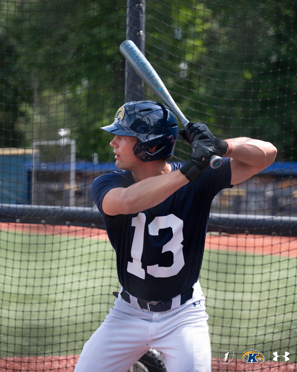 A baseball player wearing a navy jersey with the number 13 is at bat, holding a silver bat. The player's helmet features the Kent State University logo. Under Armour logos are visible on the player's pants and batting gloves. The background shows a baseball field with netting and trees.