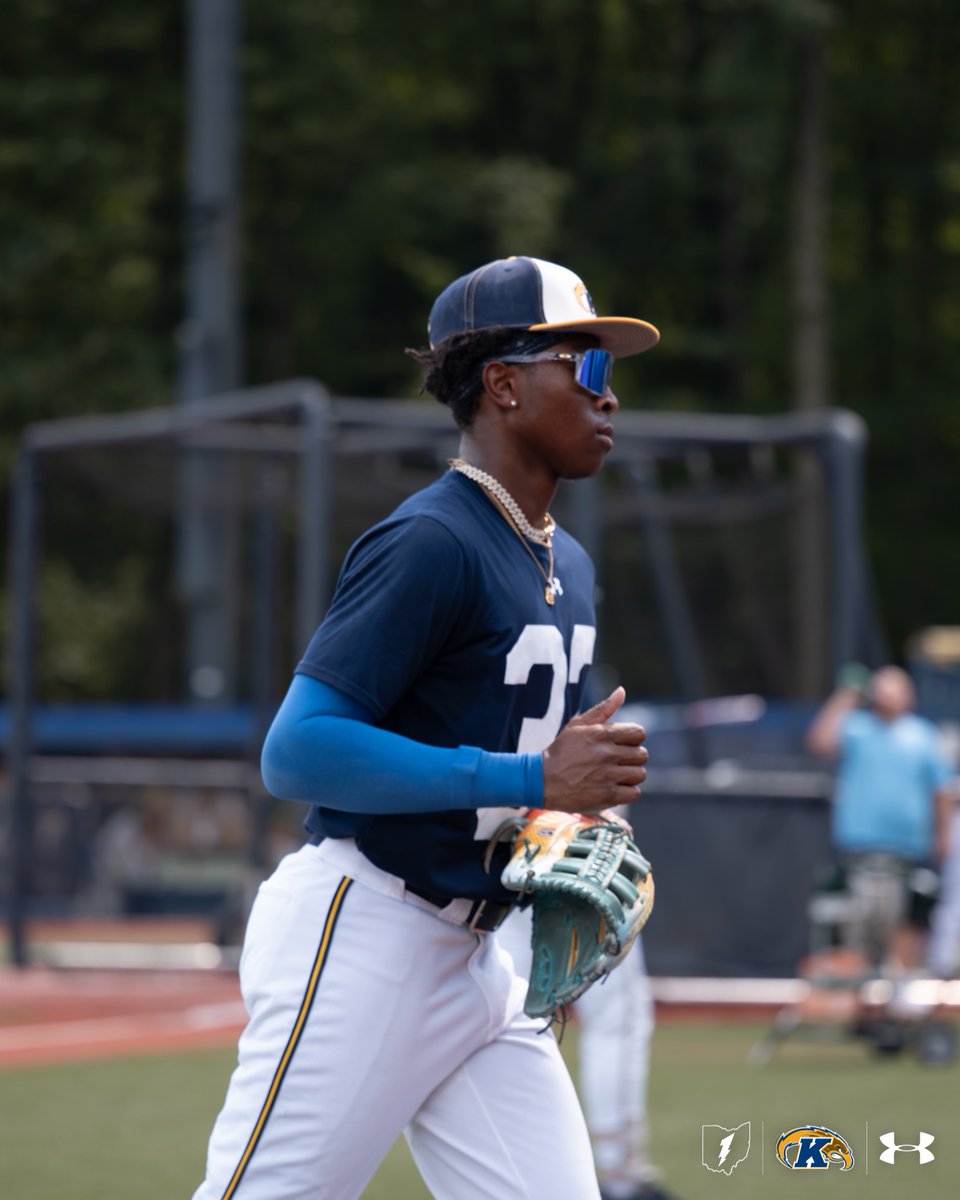 A baseball player wearing a blue and white uniform and cap, holding a glove, is on a field. The scene includes blurred spectators in the background and logos for Under Armour visible in the foreground.