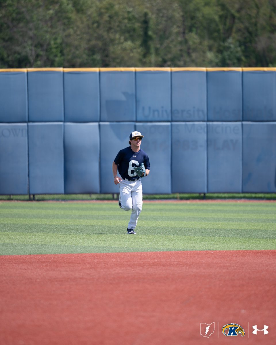 A baseball player wearing a dark shirt with the number 6 is running on a field. The background shows a blue wall with faint text from University Hospitals and Kent State University logos.