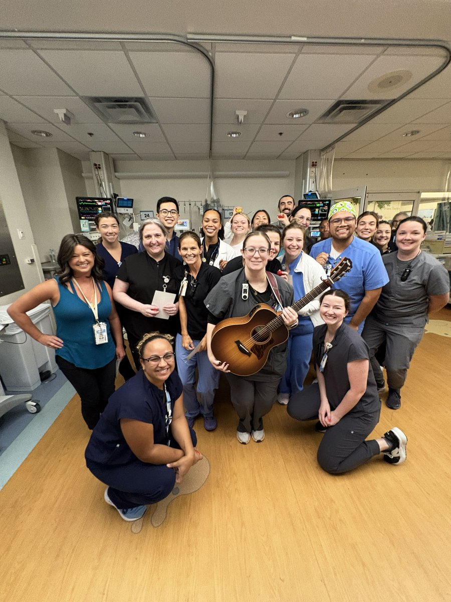 A group of hospital staff is gathered in a room, smiling at the camera. One person is holding a guitar. Medical equipment and monitors are visible in the background. The atmosphere appears cheerful and positive.