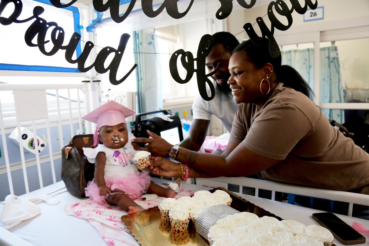A baby in a pink graduation gown is fed a cupcake in her hospital bed by her mother.